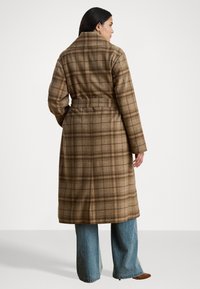 Cappotto in lana a quadri marrone con vita rifinita da una cintura, colletto ampio e lunghezza fino al polpaccio, abbinato a jeans in denim blu a gamba larga e calzature marroni.