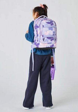 Kind mit dunklen Haaren in einem Pferdeschwanz, das eine Jeansjacke und schwarze Hose trägt, einen lilafarbenen Rucksack mit Blumenmuster auf dem Rücken hat und eine passende Wasserflasche hält.