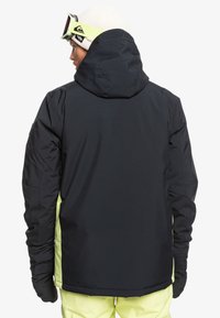 Schwarze Außensjacke mit Kapuze, mit gelben seitlichen Akzenten, strukturiertem Stoff und langen Ärmeln. Die Rückansicht konzentriert sich auf das Design und den Farbkontrast.