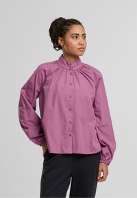 Langärmlige Bluse aus weichem, helllila Stoff. Sie verfügt über einen hohen Kragen, Raffung und eine Knopfverschlussleiste auf der Vorderseite.