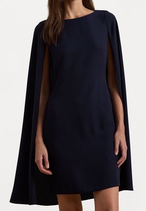 Vrouw draagt een knielange zwarte jurk met aangehechte capemouwen, staand tegen een effen lichte achtergrond.