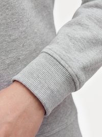 Nahaufnahme des Handgelenks einer Person, die ein hellgraues Langarmshirt mit gerippten Bündchen vor einem einfarbigen Hintergrund trägt.