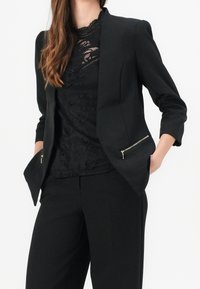 Svart blazer med skräddarsydd design, som har två zippered sidofickor, och bärs över en svart spetsblus. Slät tyg och strukturerad form.