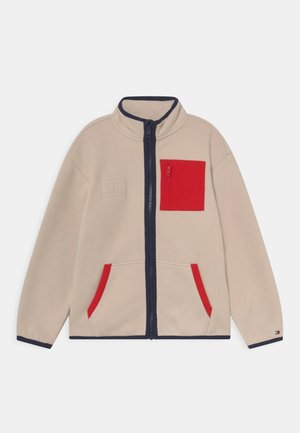 Giacca in pile beige con colletto alto, zip blu navy, tasca sul petto rossa e dettagli rossi sulle tasche. Texture morbida e vestibilità rilassata.