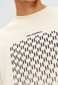 Felpa beige con un motivo geometrico nero a ripetizione e il logo "KARL LAGERFELD" sul colletto. Tessuto morbido e liscio.