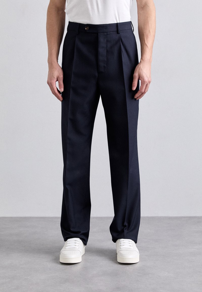Pantalons taillés bleu marine avec des plis visibles, une fermeture à bouton et des poches latérales, associés à des baskets blanches sur une surface grise.