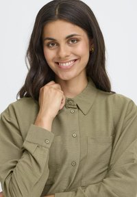 Donna sorridente con lunghi capelli scuri, che indossa orecchini a cerchio dorati e una camicia chiara oliva con bottoni, con il pugno vicino al mento.