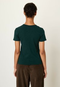 Zelena majica s kratkimi rokavi, okroglim ovratnikom in ravnim šivom na hrbtni strani. V kombinaciji z rjavimi kordnimi hlačami.