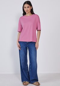 Top rosa a maniche corte con scollatura rotonda, abbinato a jeans blu a gamba larga. Il modello indossa sandali marrone chiaro. Design semplice e informale.