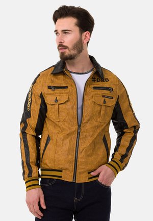 Braune Jacke mit strukturierter Textur und schwarzen Ärmeln, mit Reißverschlusstaschen und gestreiften Bündchen. Verfügt über einen Stehkragen und ein Logodesign auf der Brust.