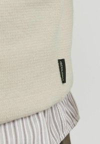 Šviesiai smėlinis megztinis su tekstūruota paviršiumi, ant kurio yra juodas etiketas su užrašu "JACK & JONES." Po juo matomas dryžuotas marškinėliai.