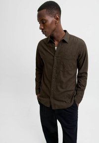 Man wearing a brown button-up shirt with a chest pocket, black pants, and gold hoop earring, looking down with hands in pockets.