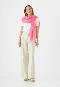 Écharpe rose drapée sur un haut blanc ; modèle portant un pantalon large crème et des sandales nudes, se tenant devant un fond blanc uni.