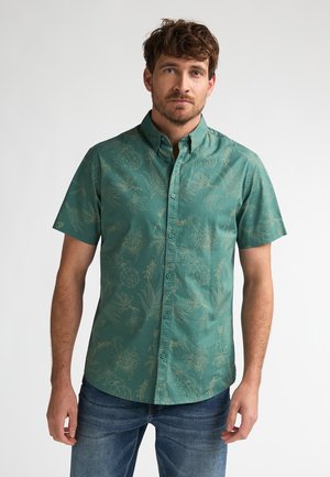 Man die een turquoise overhemd met korte mouwen en knopen en een lichte botanische print draagt en een blauwe spijkerbroek, staand tegen een effen witte achtergrond.