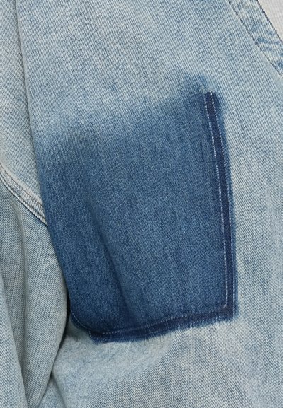 Ύφασμα τζιν με ένα πιο σκούρο μπλε ορθογώνιο κομμάτι ραμμένο πάνω σε ένα πιο ανοιχτόχρωμο μπλε φόντο τζιν, αναδεικνύοντας την υφή και τις λεπτομέρειες των ραφών.