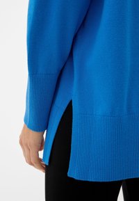 Modrý pletený svetr s žebrovanými manžetami a lemem, s bočními rozparky a měkkou texturou. Nošený přes černé legíny.