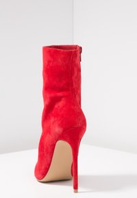 Stivali alla caviglia in suede rosso con una silhouette elegante, punta affilata, alto tacco a spillo e dettagli con cerniera laterale. Texture liscia con design minimalista.