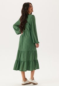 Robe verte, longueur genou, à manches longues, avec un motif en œillets, un ourlet à volants en étages et une taille cintrée. Portée avec des baskets blanches.