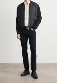 Homme portant une veste bomber matelassée noire, une chemise blanche, un pantalon slim noir, une ceinture noire et des mocassins en cuir noir, debout sur un sol uni.