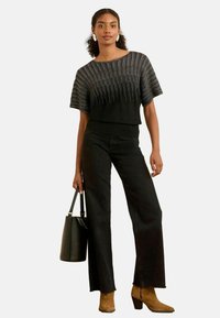 Une femme se tient avec confiance, portant un haut noir élégant et un pantalon, tenant un sac à main noir, avec des cheveux bouclés et des accessoires subtils.