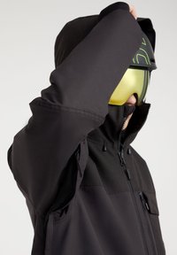 Personne portant une veste d'hiver noire avec un col zippé, levant un bras, et portant un casque noir avec des lunettes de ski jaunes.