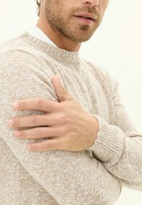 Beige knitted sweater with textured pattern, ribbed neckline and cuffs. Model holding arm, showing fabric details and skin tone.