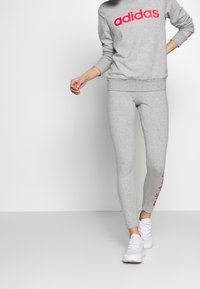 Felpa grigia con logo "adidas" rosa, abbinata a leggings grigi con branding rosa sul lato e scarpe sportive bianche.