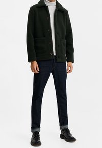 Tmavě zelená fleecová bunda s límcem, předními knoflíky a dvěma bočními kapsami, v kombinaci s tmavými džíny a černými botami.