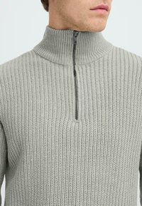 Lichtgrijze gebreide pullover met een hoge kraag en een rits tot de helft. Kenmerkt zich door een geribde structuur en verticale patronen over het geheel.