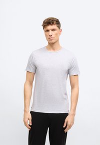 Άνδρας με σγουρά μαλλιά που φοράει ανοιχτό γκρι κοντομάνικη μπλούζα και μαύρο παντελόνι, στέκεται μπροστά σε απλό λευκό φόντο.