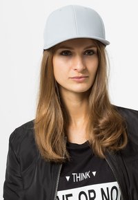 Lys grå baseballkasket lavet af tekstureret stof, med en struktureret krone og en flad skygge, parret med en sort bomberjakke og en grafisk t-shirt.