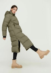 Olivengrüner, langer Daunenmantel mit Kapuze und schwarzen Akzenten, kombiniert mit hellbeigen, schnürbaren, klobigen Stiefeln und schwarzen Skinny-Jeans.