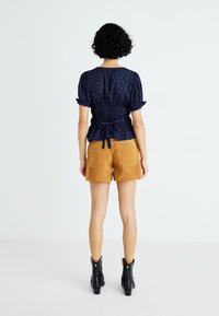 Kvinne med krøllete hår iført en marineblå topp med puffede ermer og ryggebånd, beige semsket skinnshorts og svarte ankelstøvler, stående mot en hvit bakgrunn.