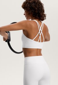 Biele športové podprsenka s prekríženým zadným dizajnom a bezšvovou textúrou, v kombinácii s bielymi legínami s vysokým pásom. Osoba drží fitness kruh.