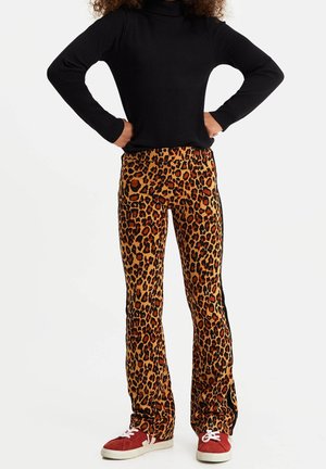 Osoba ubrana w czarną bluzkę z długim rękawem, spodnie w panterkę z czarnymi bocznymi paskami oraz czerwone trampki, stojąca z rękami na biodrach.