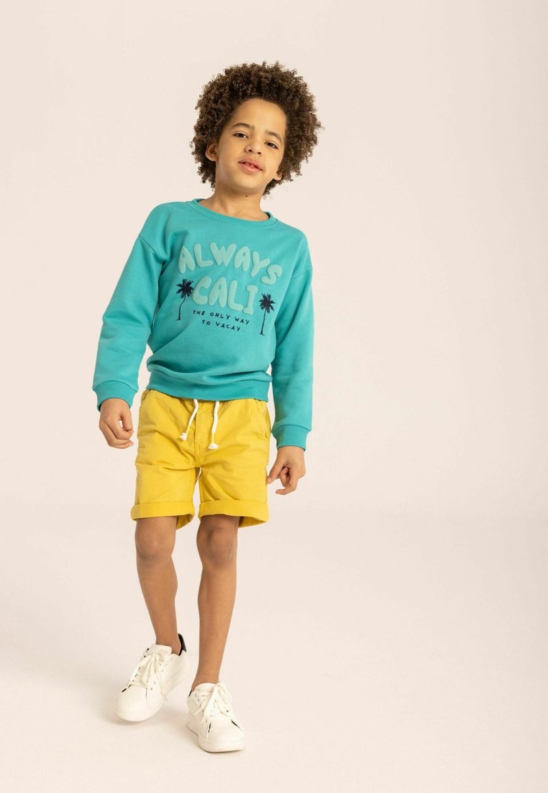 Turkos sweatshirt med tryck "ALWAYS CALI" och palmträd; gula shorts, vita sneakers. Avslappnad outfit för aktivt bruk.