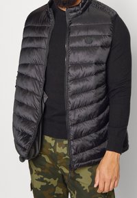 Svart puffer-väst med horisontell quiltad design, dragkedja, hög krage och en logotyp på bröstet. Bärs över en svart långärmad topp.