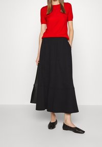Top rosso a maniche corte abbinato a una gonna nera al ginocchio con un design svasato. Scarpe nere basse completano l'outfit.
