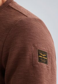 Smešasta smeđa vesta s reljefnim ovratnikom i kantama na ramenima, s kvadratnom crnom etiketom sa zlatnim natpisom na rukavu.