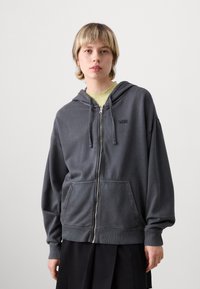 Tmavě šedá mikina s kapucí na zip, se stahovací šňůrkou, předními kapsami a mírně oversize střihem. Na hrudi je malá černá značka.