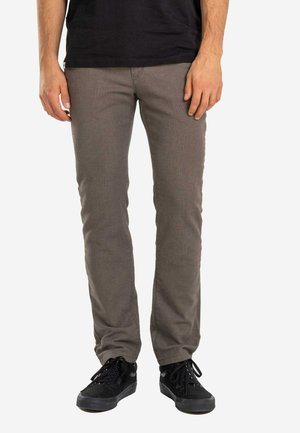 Mann trägt schlank geschnittene braune Hose und schwarze Sneakers, steht vor einem schlichten weißen Hintergrund.