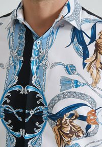 Chemise à manches courtes avec un mélange de motifs floraux bleus sur fond blanc, dotée d'un panneau noir et de détails de boutons. Texture de tissu lisse.