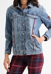 Jeansjacka med ljusblå tvätt, röda sidsömmar, knäppning med knappar, två bröstfickor och ett stjärnmärke på höger ärm.