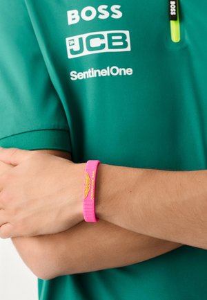 Groen poloshirt met korte mouwen en witte logo's. Een felroze siliconen polsbandje toont "FORMULA ONE TEAM" in geel.