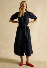 Tumedat sinise puuvillane midi kleit, mis on varustatud eest kinnituvate nööpidega, puffvarrukate, stringiga talje ja astmeline seelik; koos punaste lamedate kingadega.