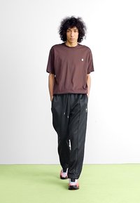 Brun kortærmet T-shirt parret med sorte sweatpants. Bukserne har snøre og diskret branding. Modellen har farverige sneakers på.
