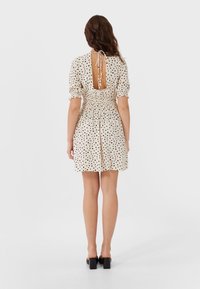 Cremefarbenes Kleid mit schwarzen Polka-Dots, kurzen Puffärmeln und einem offenen Rücken, der mit einem Band gesichert ist. Es verfügt über eine geraffte Taille und einen ausgestellten Rock.
