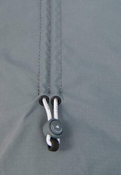 Γκρι ύφασμα με λεπτή ανάγλυφη υφή ραβδώσεων, που διαθέτει δύο μαύρους μηχανισμούς σύσφιξης κορδονιών και λευκά κορδόνια. Η ορατή ραφή αναδεικνύει την ανθεκτικότητα.