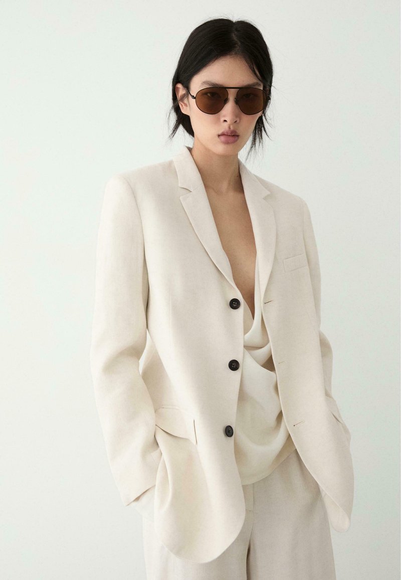 Osoba ubrana w oversize'owy kremowy garnitur z czarnymi guzikami, luźne spodnie w tym samym kolorze, bluzkę z głębokim dekoltem w kształcie litery V oraz brązowe okulary przeciwsłoneczne, stojąca na jednolitym tle.