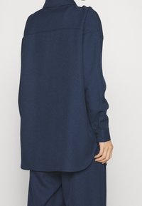 Mörkblå långärmad skjorta, avslappnad passform, mjukt material, rundad fåll, detalj med rygghalssöm, knappmanschett, synliga sömnadsdetaljer.